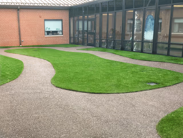 Outdoor courtyard area with green grass patches and winding paved pathways, surrounded by brick walls and large glass windows of the building.