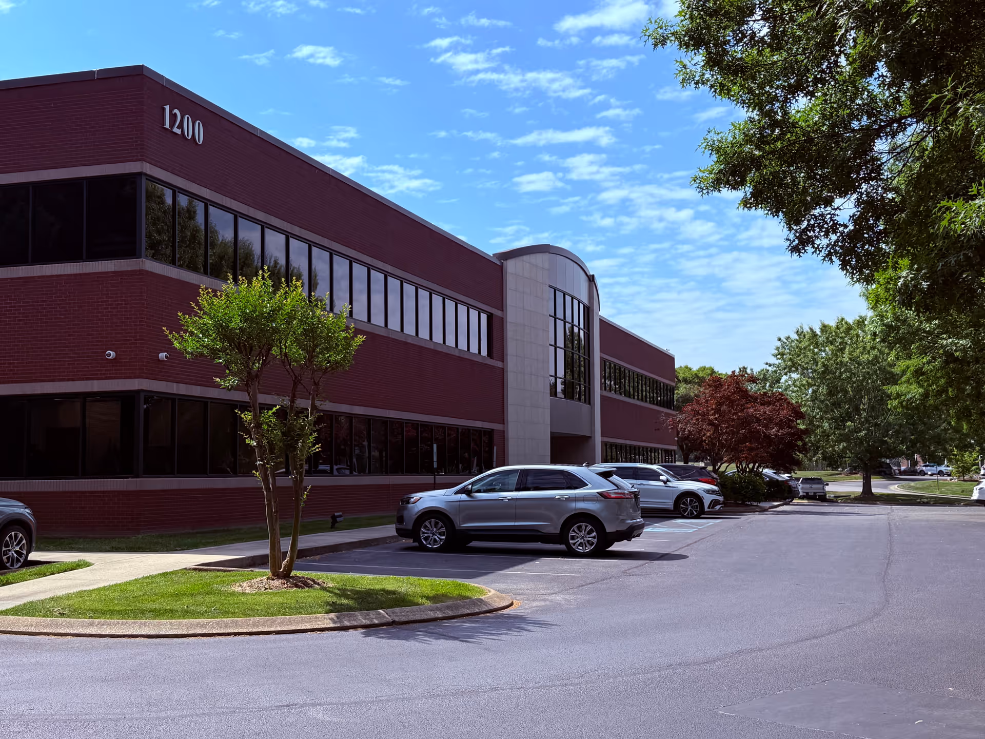 Two-story brick building with large windows, parking, and trees.
