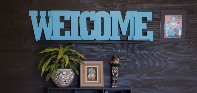 Interior wall with a blue WELCOME sign, potted plant, framed picture, and figurine on a shelf.