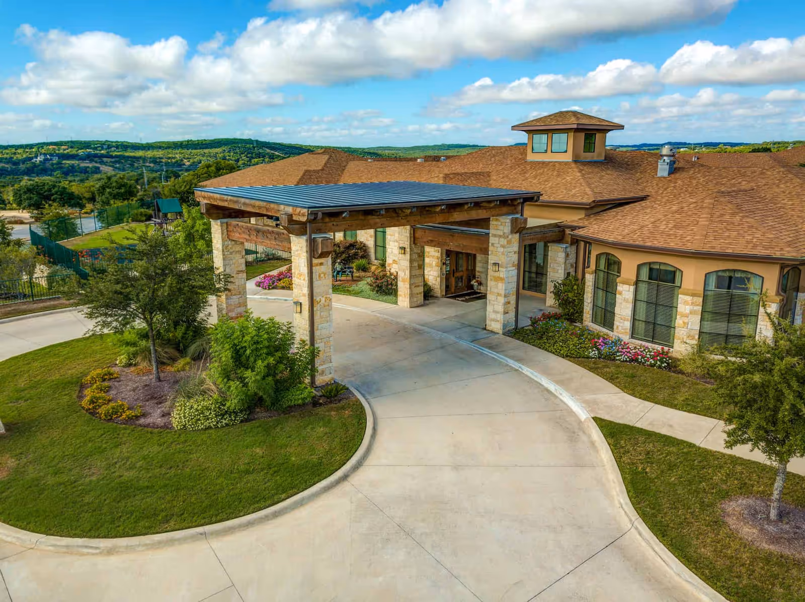 Covered porte-cochere and curved driveway leading to the entrance of a single-story stone and stucco building surrounded by landscaped lawns and distant hills.