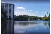 Exterior view of a multi-story building beside a calm river with trees and blue sky.
