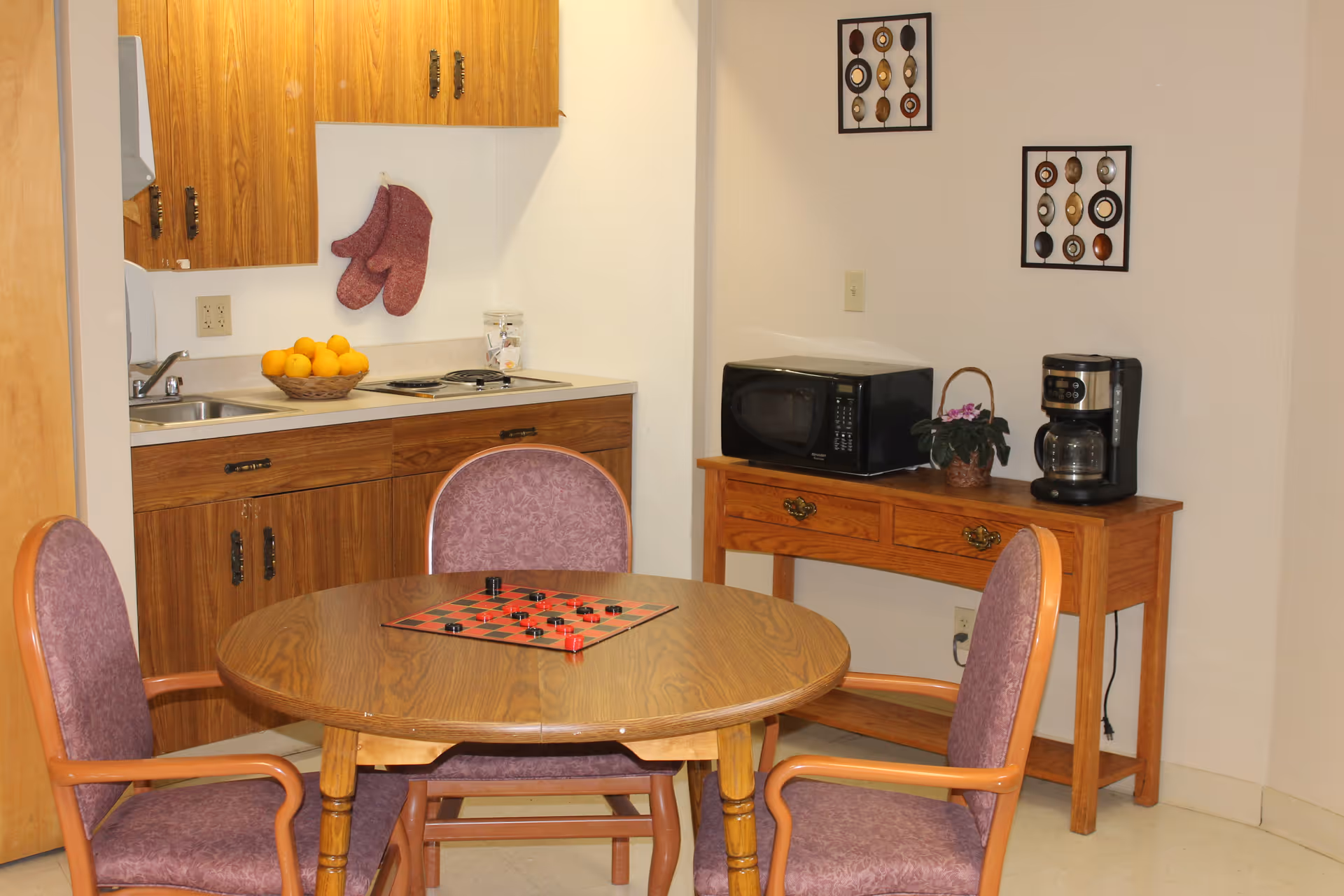 A small kitchenette area with wooden cabinets, a sink, and a stovetop. A basket of oranges sits on the counter. In front of the kitchenette is a round wooden table with a checkers game set up and three purple upholstered chairs around it. To the right, there is a wooden side table with a microwave, a coffee maker, and a small potted plant. Two decorative wall hangings are displayed above the side table.