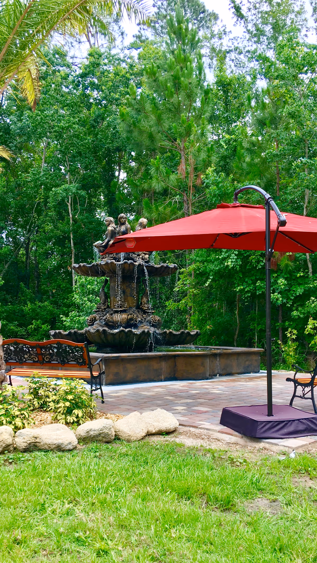 Outdoor patio area with a large decorative water fountain featuring sculptures, surrounded by green trees. There is a red umbrella providing shade over a seating area with benches and chairs on a paved surface.