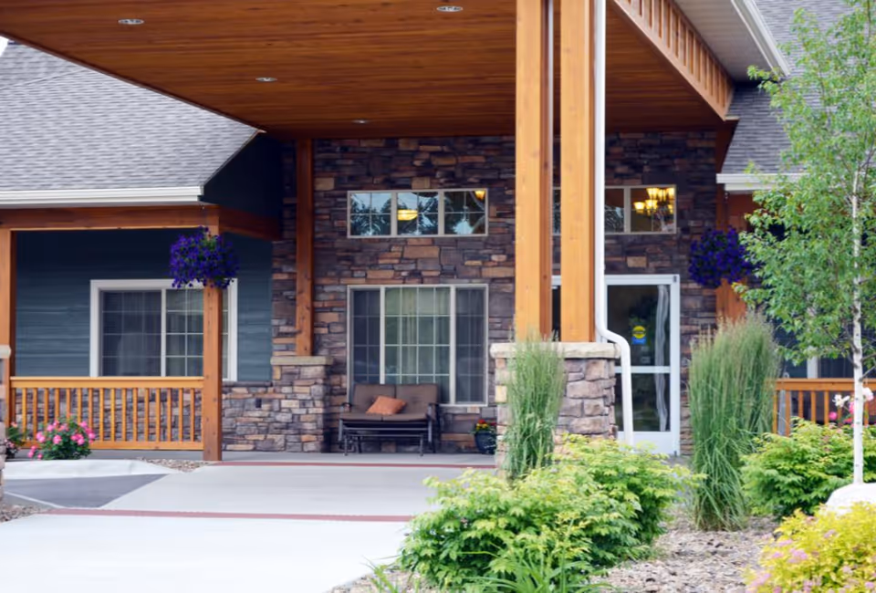 Covered entrance of an assisted living building with stone facade, wooden columns, seating on the porch, and landscaped greenery.