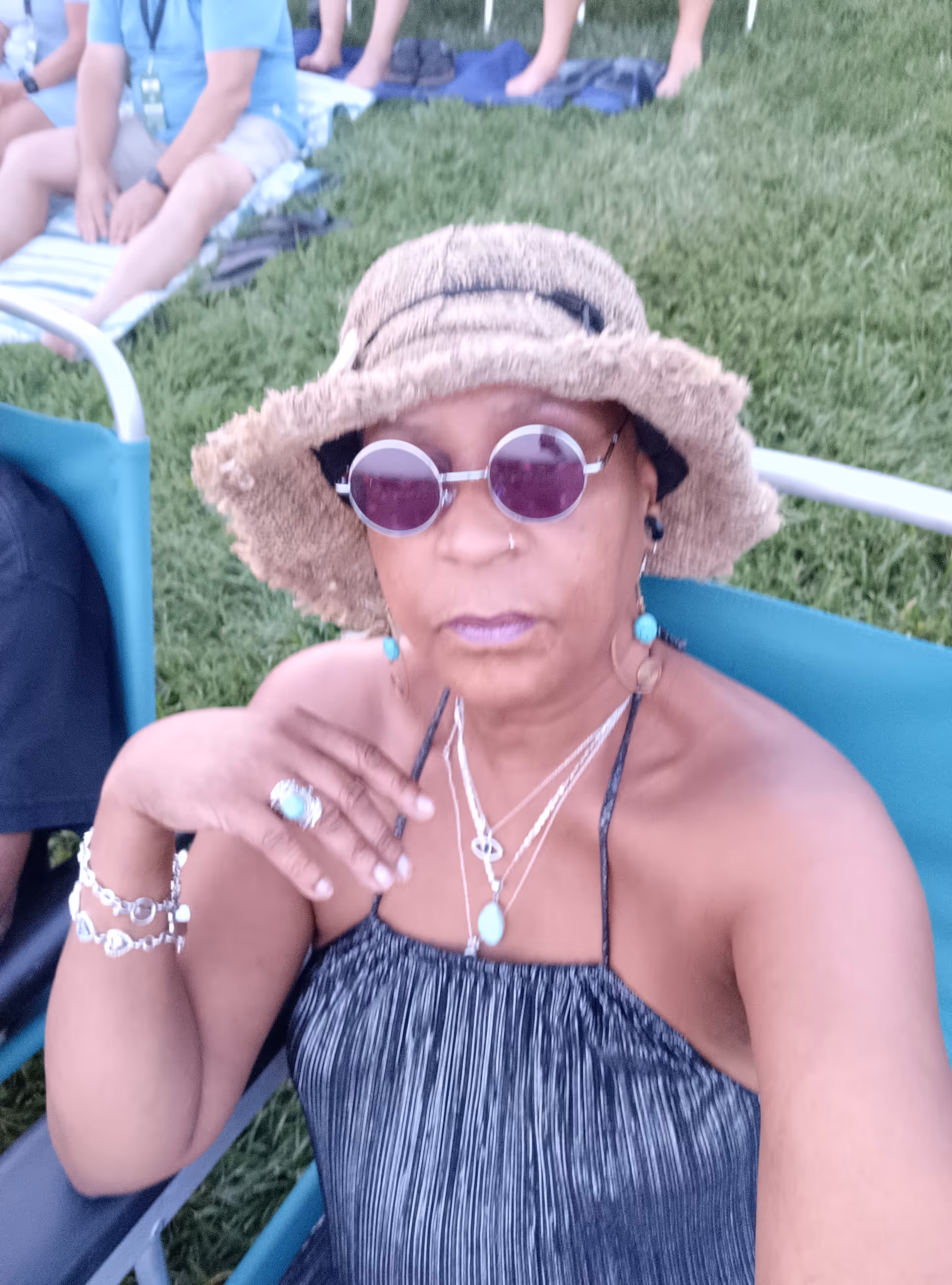 Senior woman wearing straw hat and purple sunglasses sits outdoors on grass with others nearby.