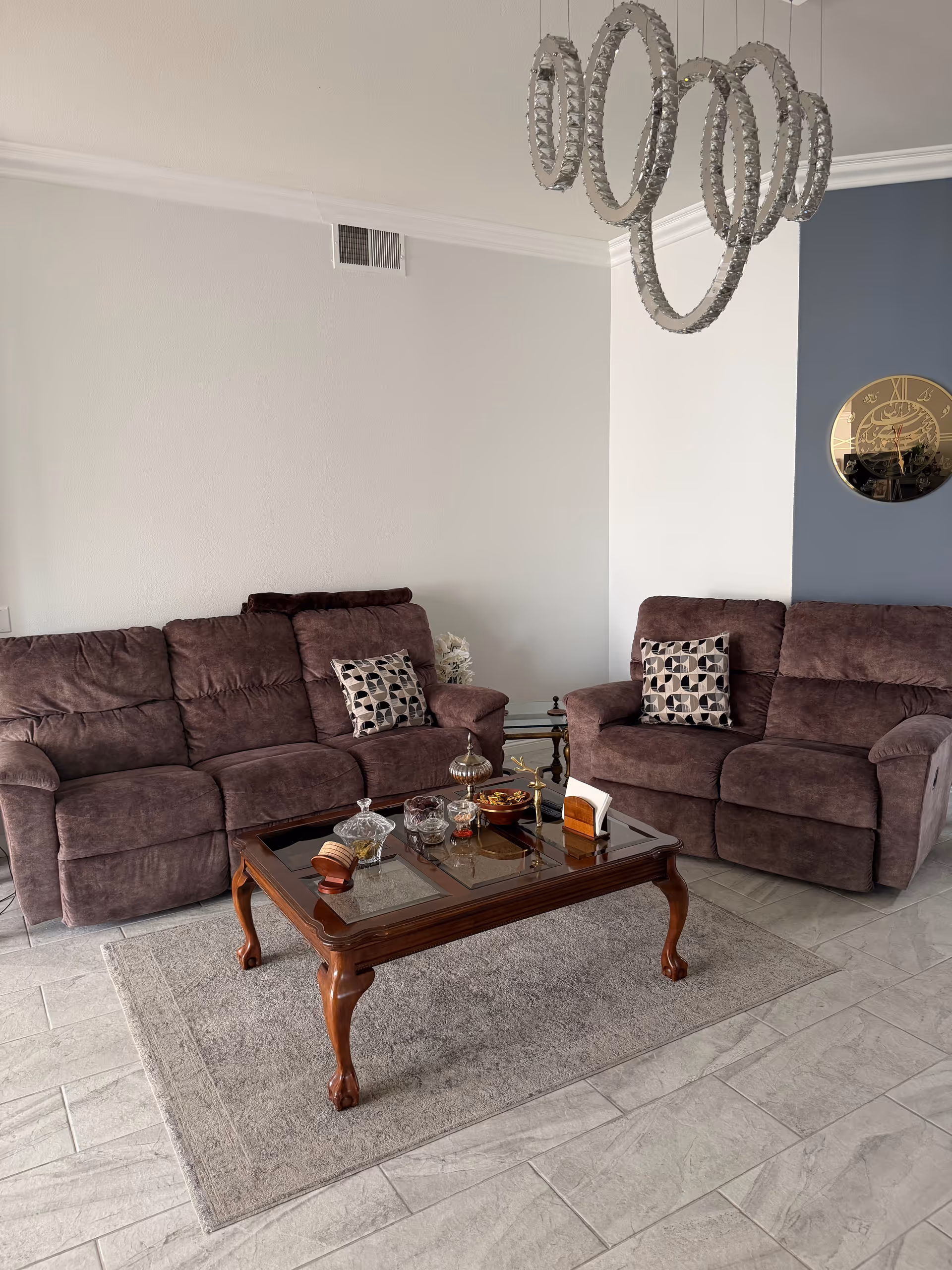 A cozy living room with two brown couches, cushions, glass coffee table, and chandelier.
