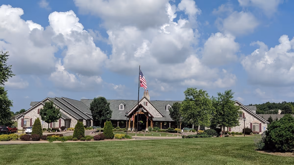 Front view of a single-story stone-clad senior living building with an American flag, landscaped lawn, and a partly cloudy sky.