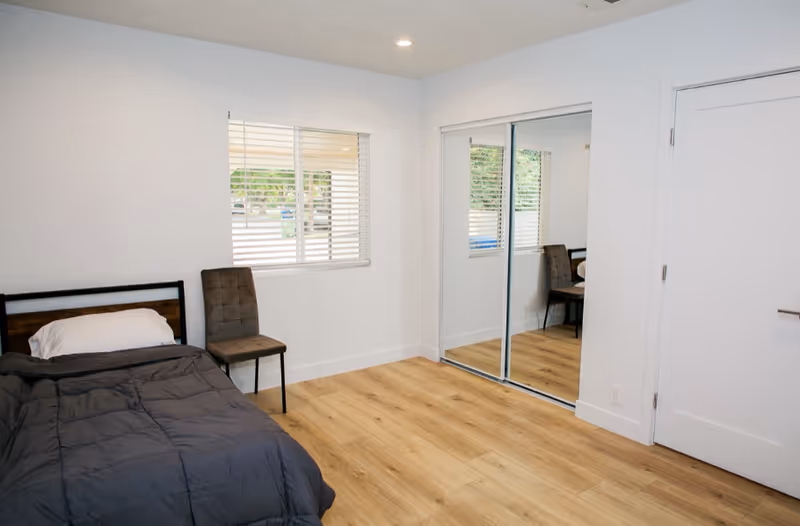 Bedroom in senior living facility with a bed, chair, window, and mirrored closet.