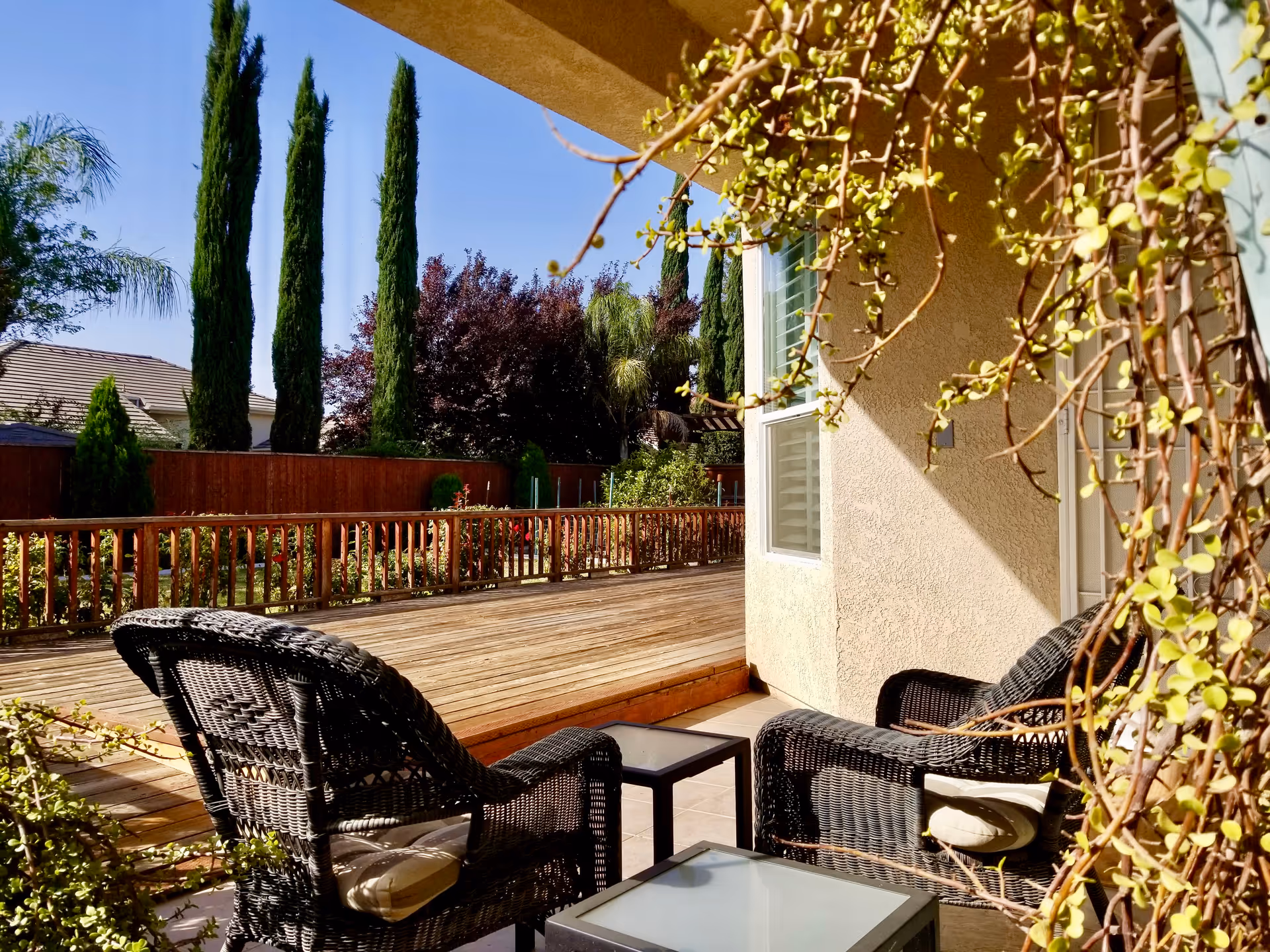Sunny outdoor patio with wicker chairs, glass table, and wooden deck beside a beige villa.