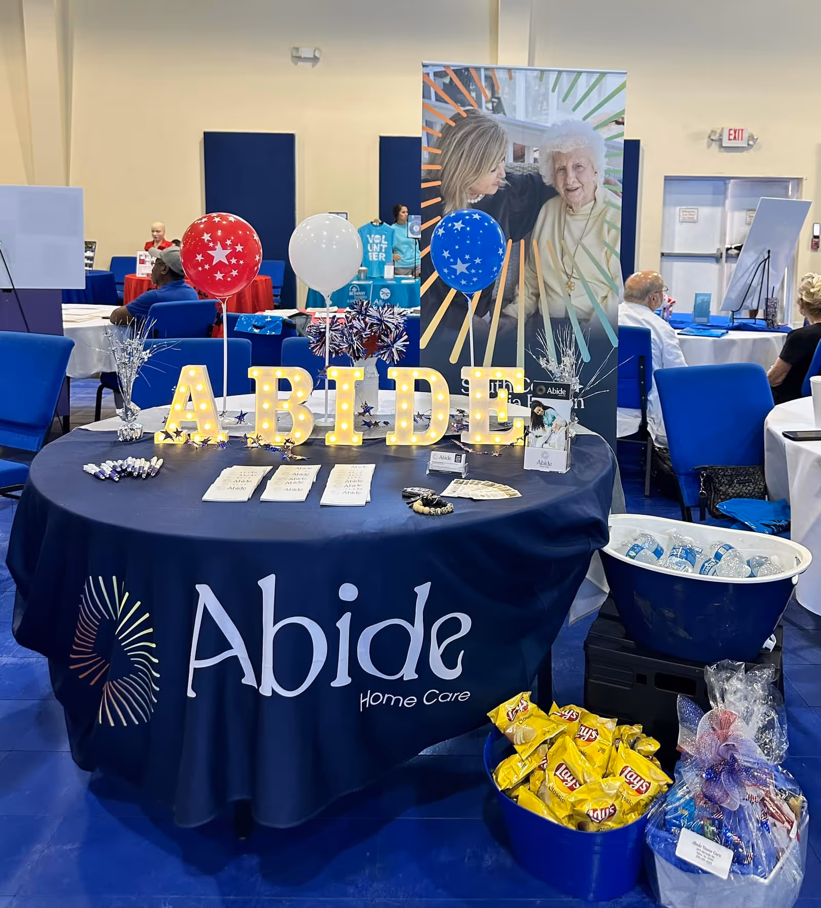 Round exhibition table with Abide Home Care branding, illuminated ABIDE letters, balloons, and a banner.