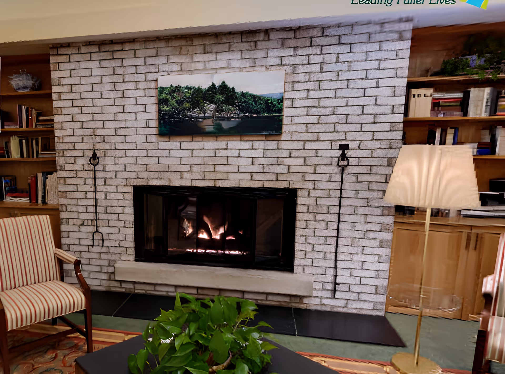 A cozy living room area featuring a lit fireplace set in a white brick wall. Above the fireplace is a landscape painting of a forested area with a body of water. On either side of the fireplace are built-in wooden bookshelves filled with books and decorative items. In front of the fireplace is a coffee table with a green leafy plant. To the left is a striped armchair, and to the right is a tall floor lamp with a beige shade.