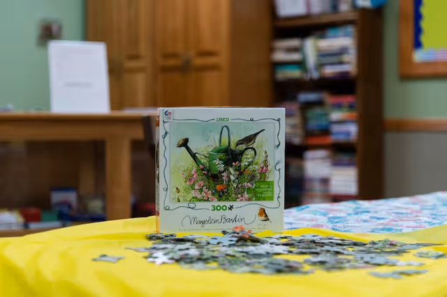 A 300-piece jigsaw puzzle box featuring a watering can and flowers is placed on a yellow tablecloth with scattered puzzle pieces in front. In the background, there is a wooden cabinet, a bookshelf filled with books, and a bulletin board on the wall.