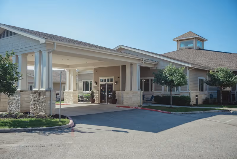 Front entrance of a senior living facility with a covered porte-cochère, stone-clad columns, and a landscaped driveway.