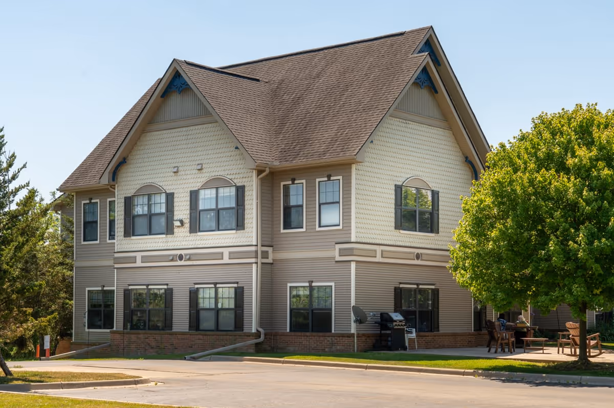 Exterior view of a two-story senior living facility building with beige siding, multiple windows with black shutters, and a brown shingled roof. There is a paved driveway in front and a green tree on the right side with outdoor seating and a grill visible under the tree.