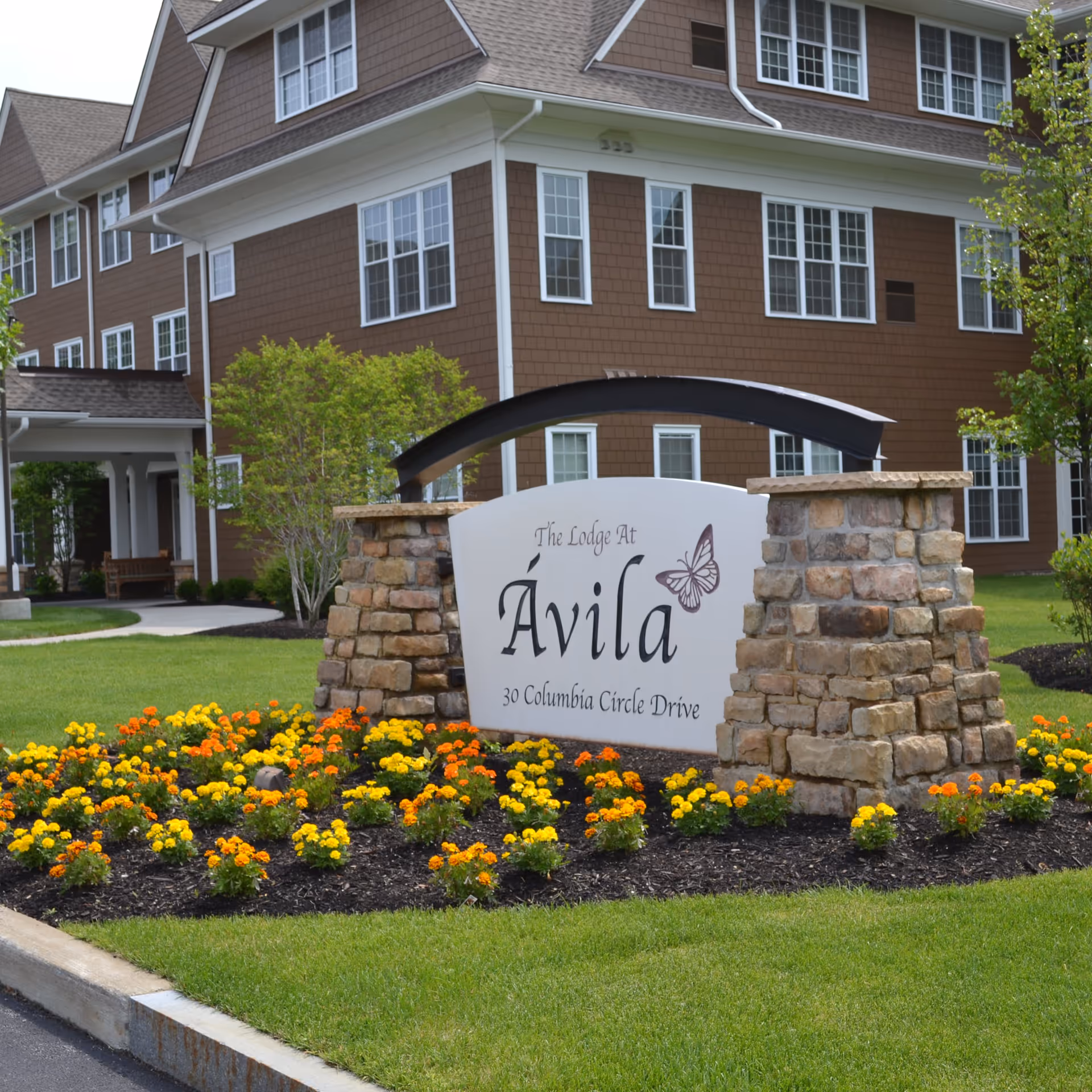 The exterior of a brown multi-story building with white-trimmed windows, surrounded by green grass and colorful orange and yellow flowers. In front of the building is a stone sign that reads 'The Lodge At Avila, 30 Columbia Circle Drive' with a butterfly graphic.