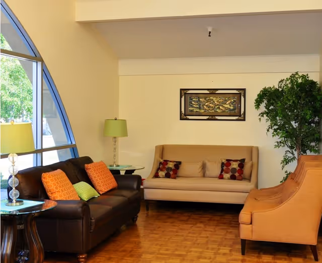 Seating area in a senior living room with leather and fabric sofas, accent chairs, colorful pillows, lamps, a framed artwork, and a large arched window.