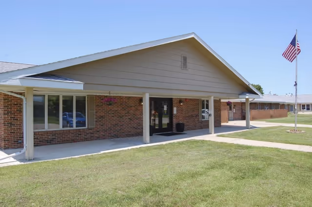 Exterior view of a single-story brick building with a covered entrance supported by columns, a large window, and an American flag on a flagpole on a grassy lawn under a clear blue sky.