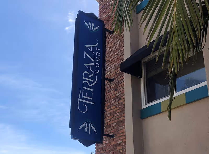 Vertical blue sign that reads "Terraza Court" mounted on a brick building facade with a palm frond nearby.