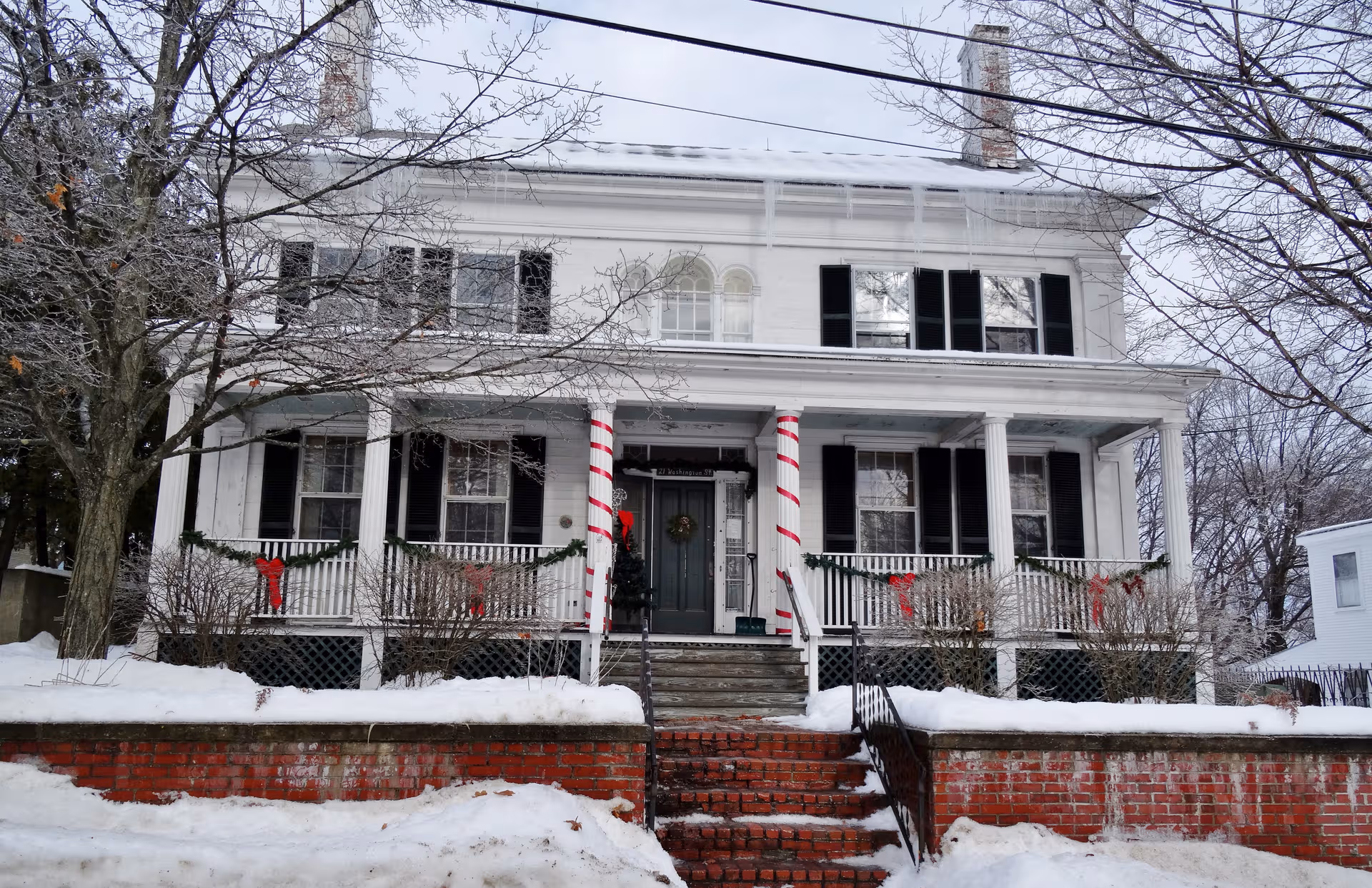 Snow-covered, two-story white house with a decorated front porch and striped candy-cane columns.
