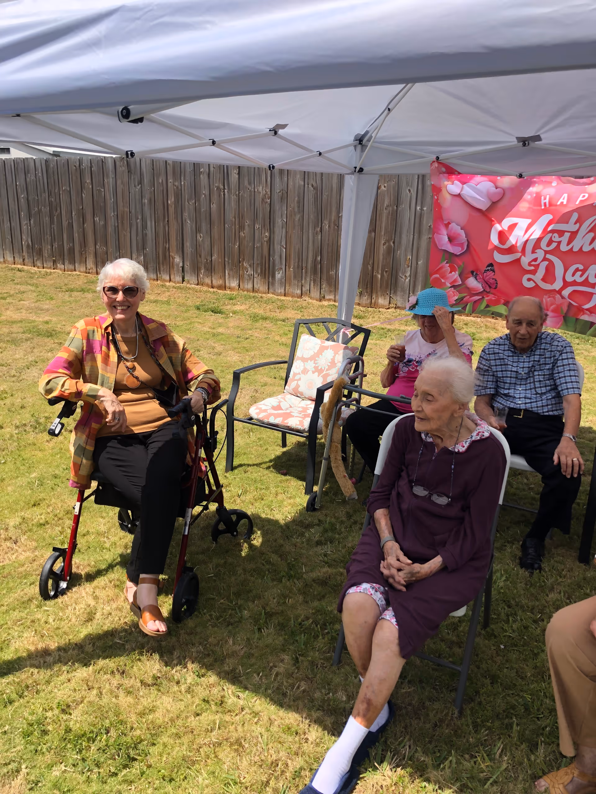 Four elderly people sitting outdoors under a white canopy tent on a grassy lawn. One woman with white hair and sunglasses is sitting on a red walker, smiling. Another elderly woman with white hair is seated on a chair wearing a purple dress. Behind them, a man and another woman are seated on chairs near a wooden fence. A pink banner with floral designs and the words 'Happy Mother's Day' is hanging on the canopy.