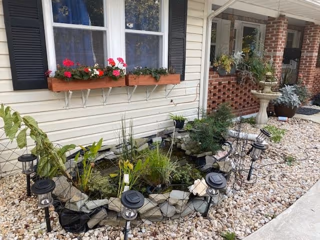 Stone-lined garden pond and pebble landscaping in front of a house with window flower boxes and a brick porch.