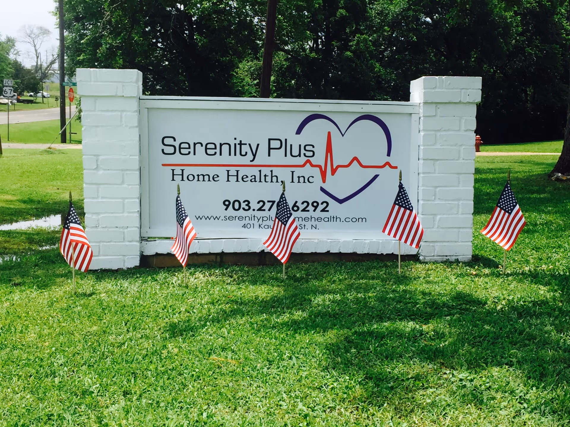 A white brick sign for Serenity Plus Home Health sits on a grassy lawn with flags.