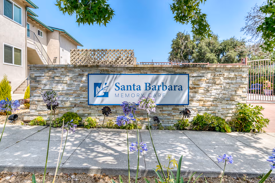 Stone-faced building entrance with Santa Barbara Memory Care sign, surrounded by landscaping and a metal gate.