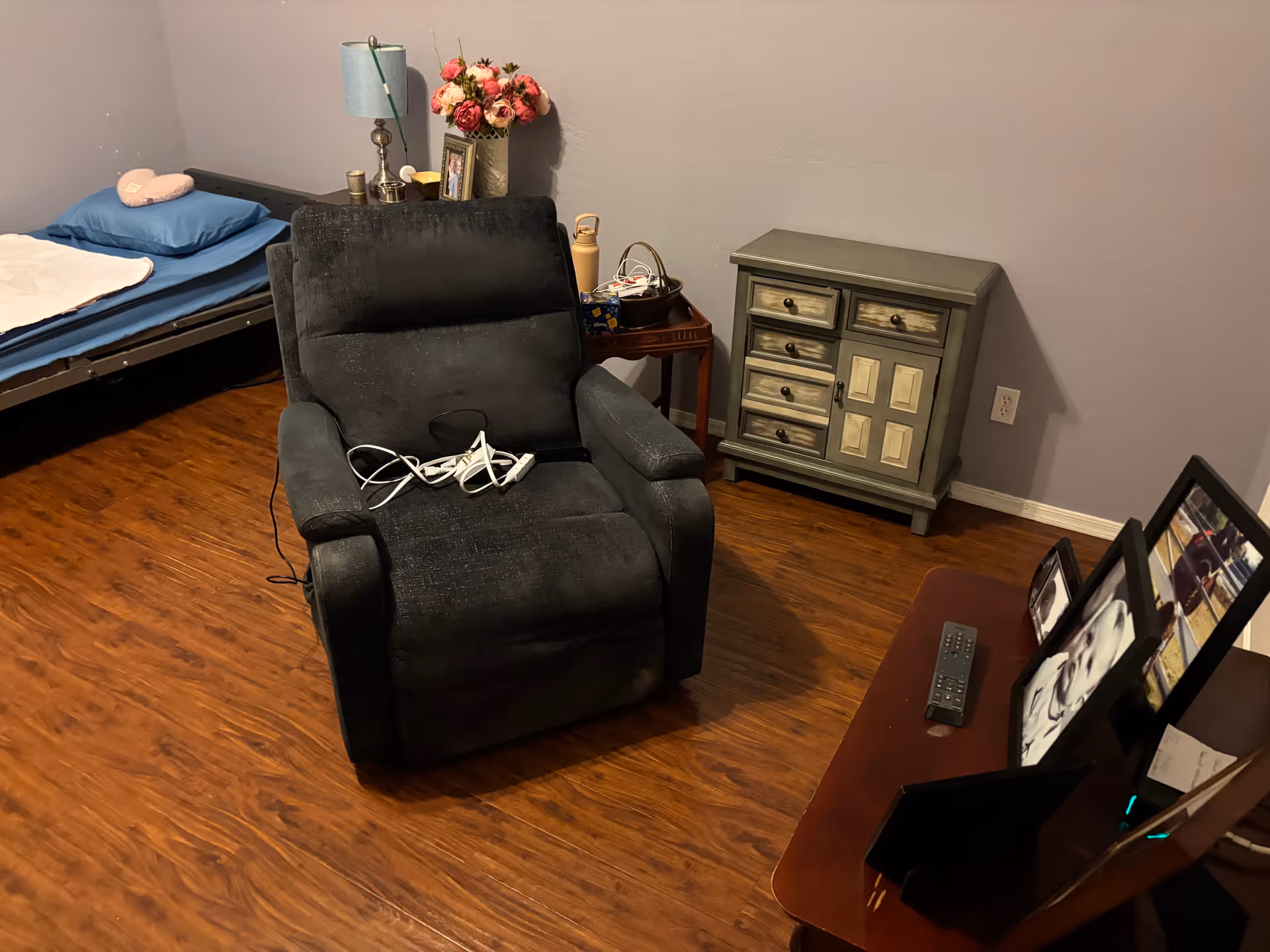 Cozy bedroom in a senior care facility with bed, recliner, dresser, and framed photos.