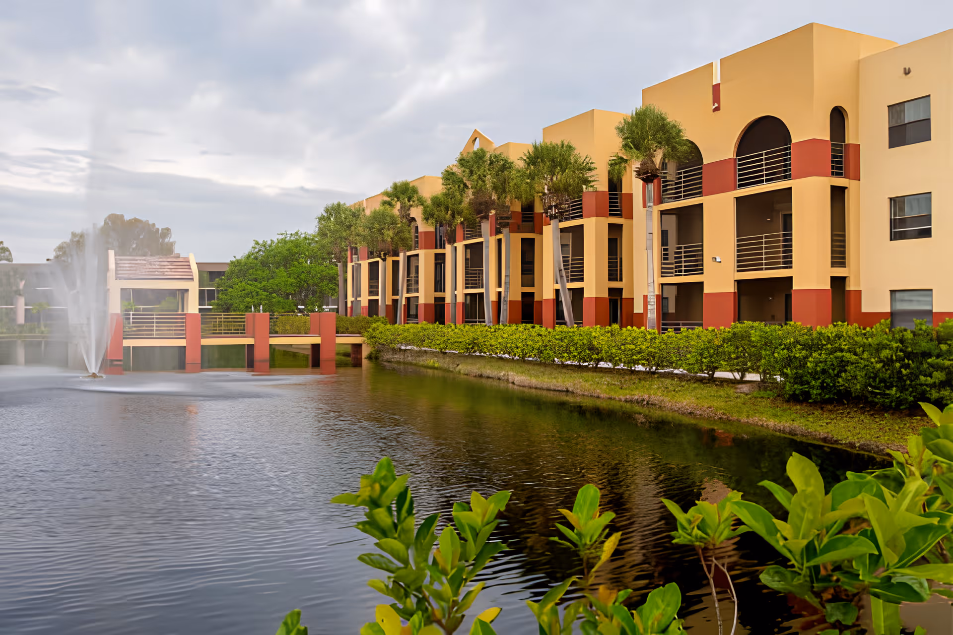 Three-story beige-and-red assisted living building with balconies overlooking a landscaped pond and fountain.