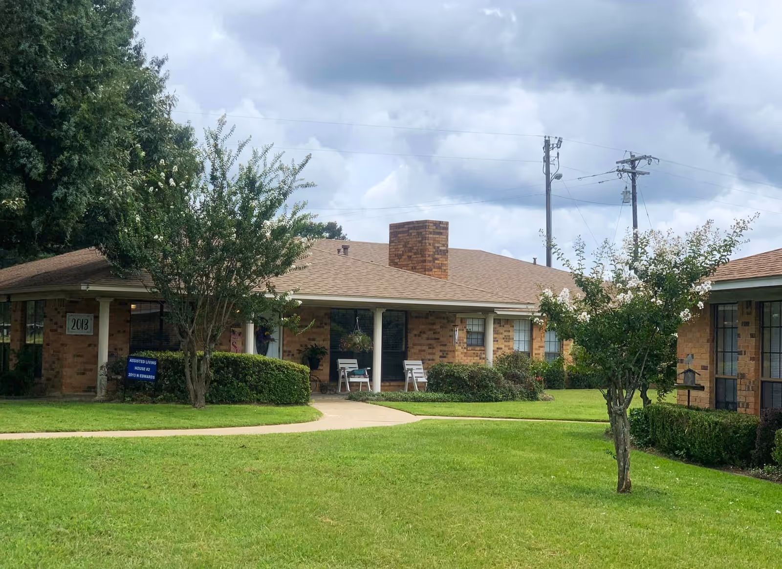 Exterior view of a single-story brick building with a covered porch, two white chairs, and a chimney. The building is surrounded by green grass, bushes, and small trees under a cloudy sky.