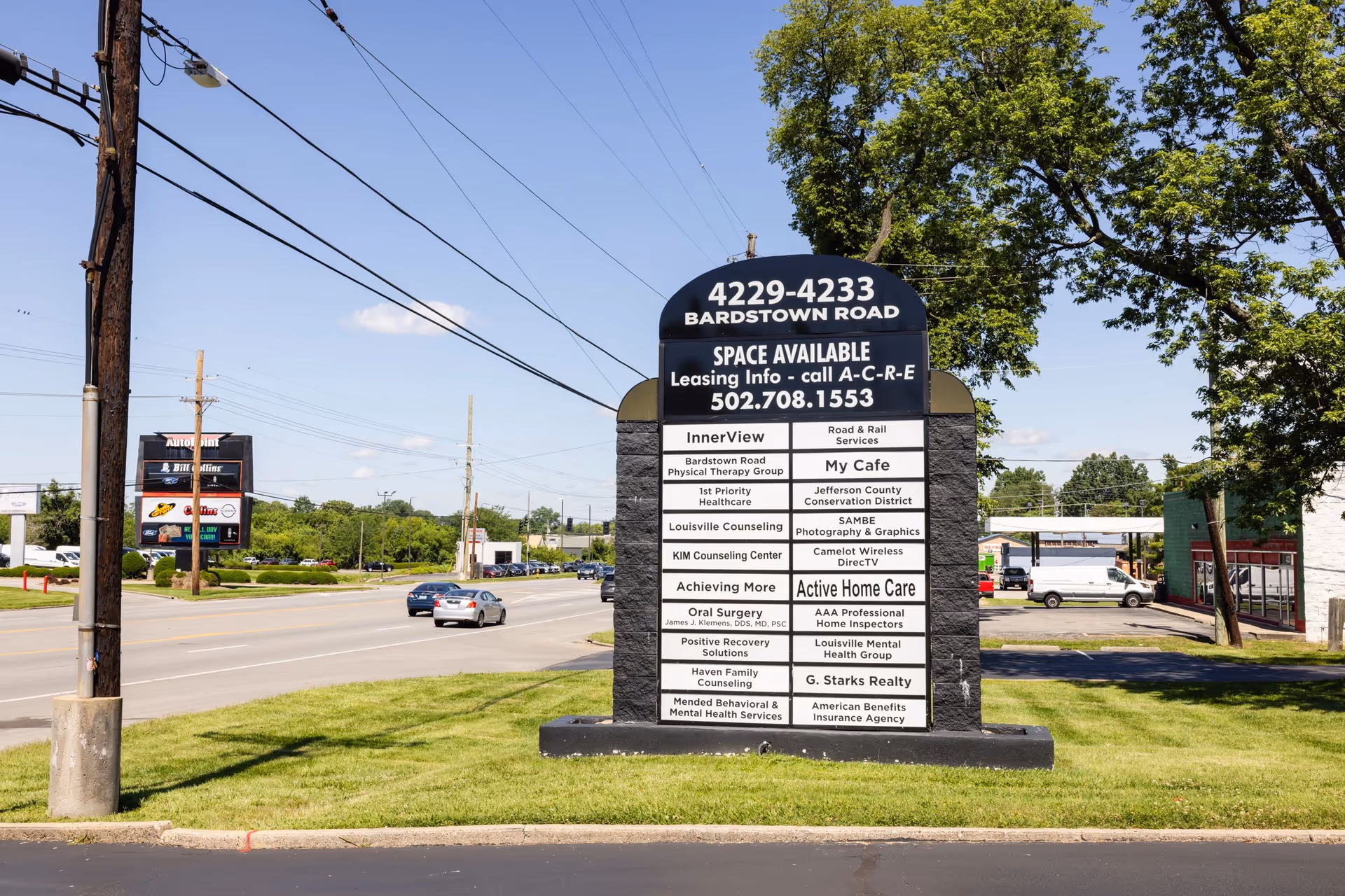 Tall roadside sign with business listings on a grassy median beside a busy road.