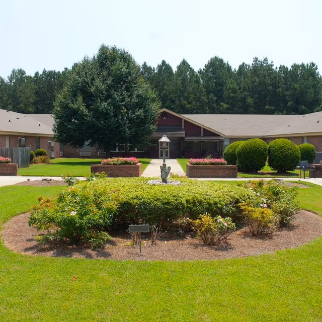 A landscaped garden area with trimmed bushes and a small statue in the center, surrounded by a circular patch of grass. Behind the garden is a single-story brick building with a peaked roof, large tree, and neatly trimmed shrubs. The scene is set against a backdrop of tall pine trees under a clear sky.
