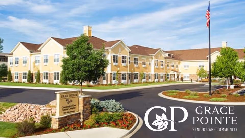 Exterior view of Grace Pointe Senior Care Community building with a well-maintained garden, a tree, and a sign displaying the facility's name. The building is two stories with multiple windows and a brown roof under a partly cloudy sky.
