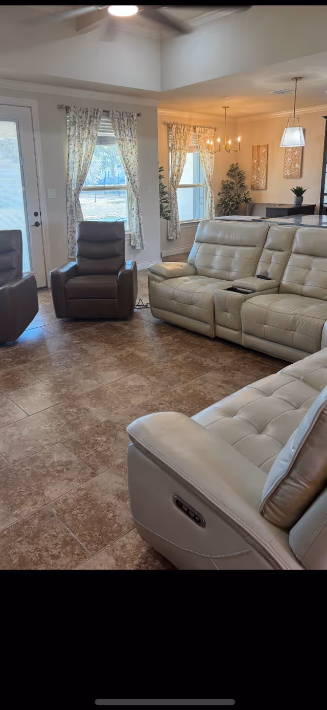 Bright living room with beige leather seating, windows with floral curtains, and a dining area.