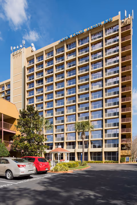 Exterior view of a multi-story senior living facility building with numerous windows, a small gazebo, palm trees, and parked cars in front under a clear blue sky.