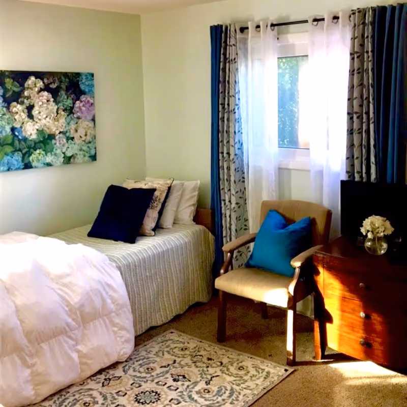 Sunlit bedroom with a neatly made bed, chair, dresser, window, and floral painting.