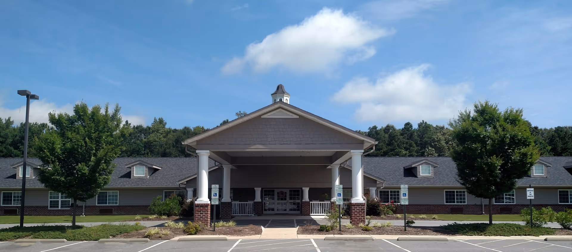 Front exterior of a single-story senior living building with a covered porte-cochère entrance, parking spaces, and trees under a blue sky.