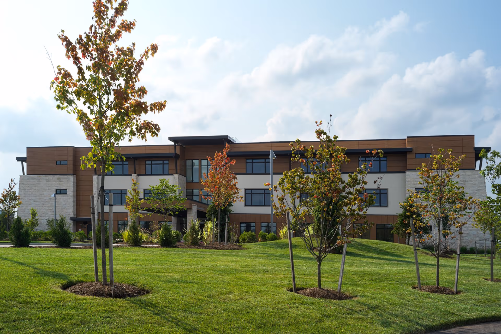 Exterior view of a modern three-story building with a mix of stone and wood paneling, surrounded by a well-maintained lawn and young trees with some autumn-colored leaves under a partly cloudy sky.