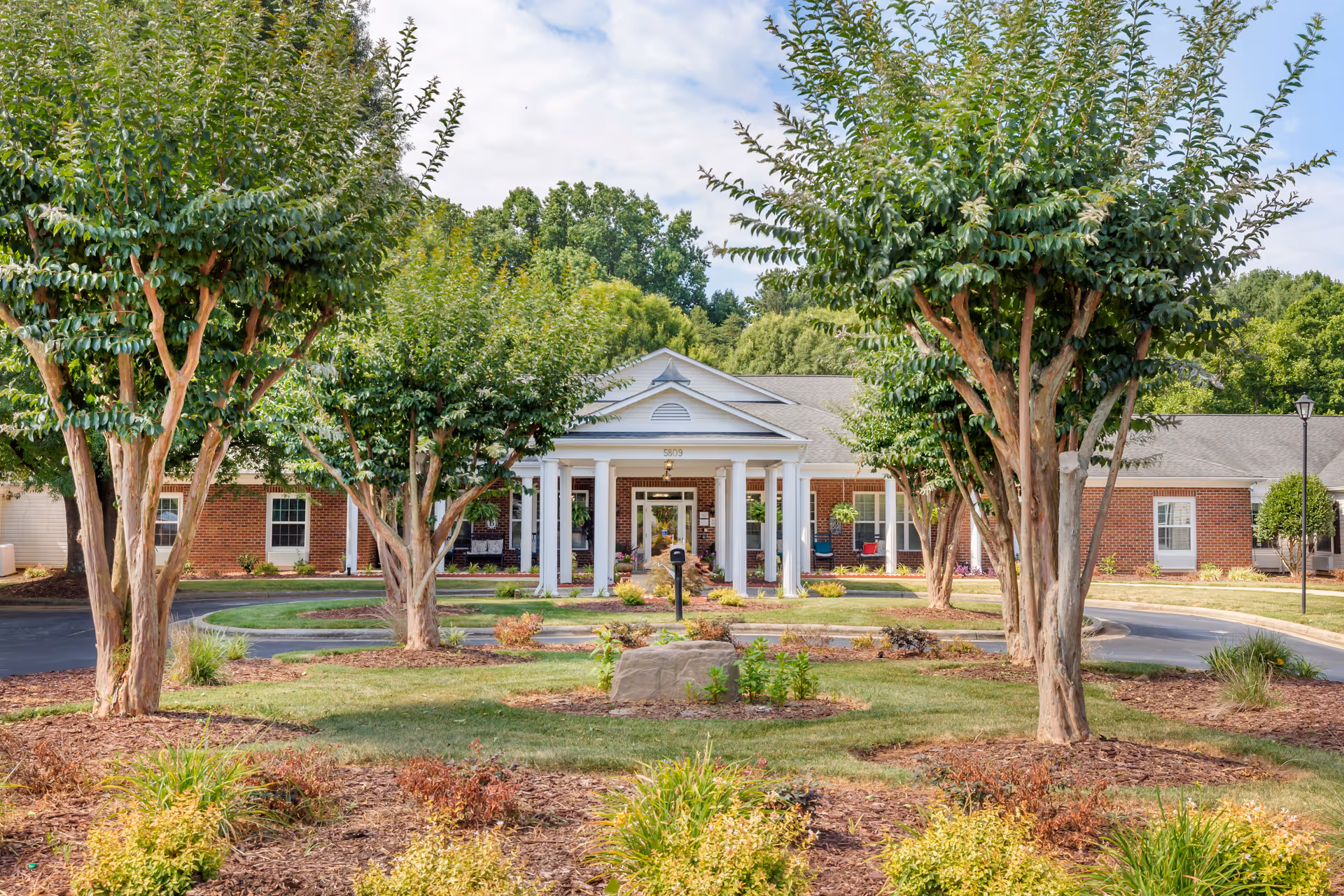 Front exterior view of Brookdale Northwest Greensboro facility showing a brick building with white columns at the entrance, surrounded by neatly landscaped gardens and trees under a partly cloudy sky.