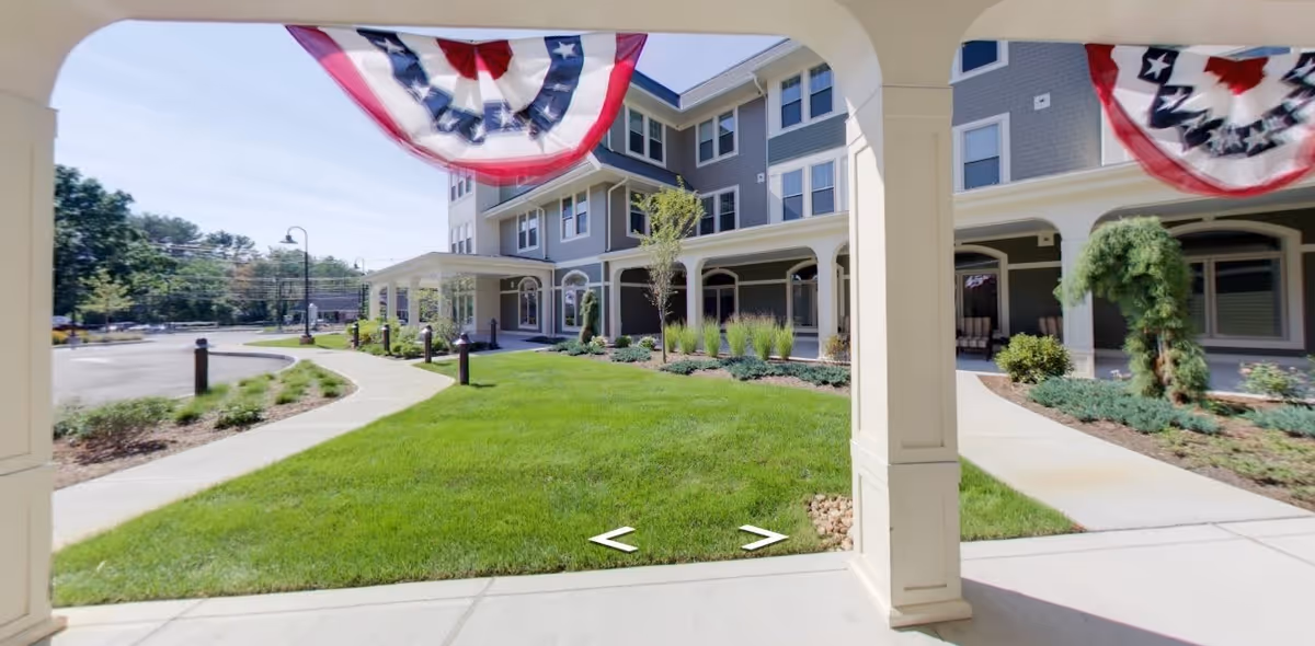 View of the exterior front entrance of a senior living facility named The Residence at Five Corners, showing a well-maintained lawn, paved walkways, and patriotic bunting decorations hanging from the covered porch area.