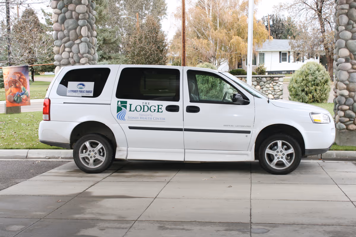 A white van parked on a driveway with logos on the side reading 'The Lodge Managed by Sidney Health Center' and 'Community Public Transit'. The background shows stone pillars, trees, and a residential area.