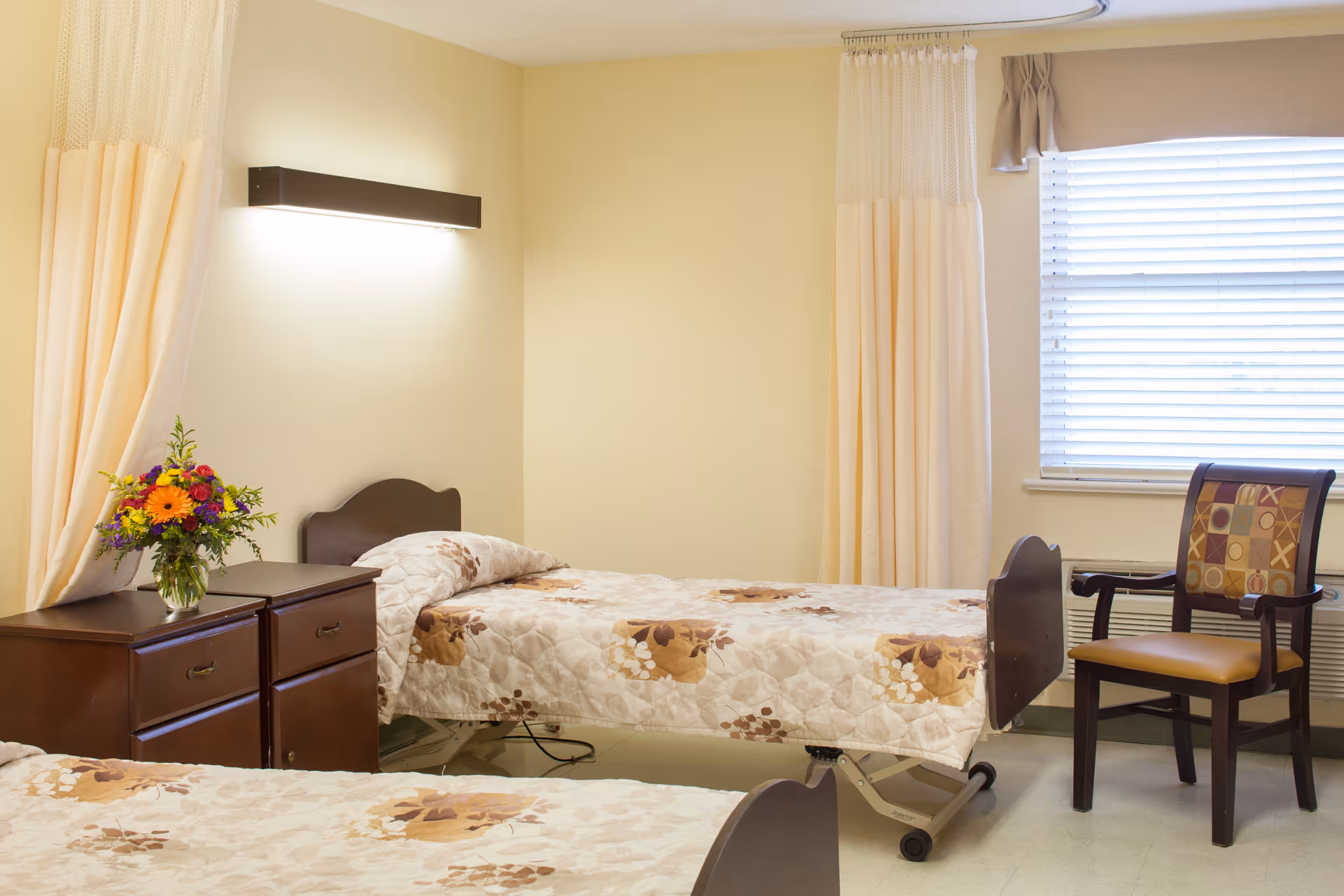 A simple, clean bedroom in a senior living facility with two single beds covered in floral patterned bedspreads. There is a wooden nightstand with a vase of colorful flowers between the beds. A wooden chair with a patterned cushion is placed near a window with closed blinds and beige curtains. The walls are painted light yellow and a wall-mounted light fixture is above one of the beds.