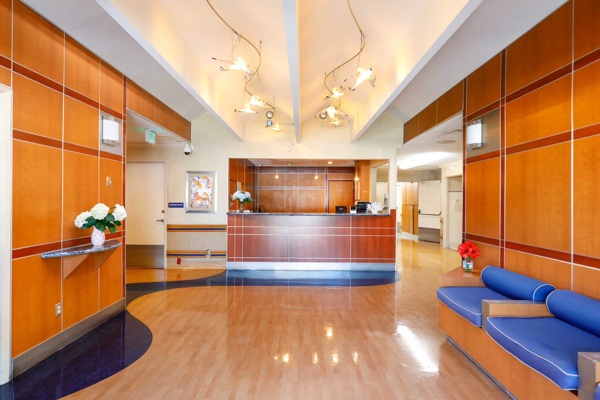 Reception lobby with a wooden front desk, blue bench seating, and decorative ceiling lights.