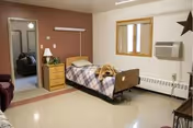 Resident room with adjustable hospital bed, wooden dresser, window, and a dog resting on the bed.