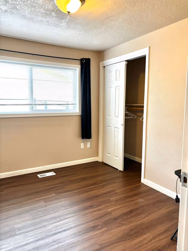 Empty bedroom with wood floor, window, dark curtain, and open closet.