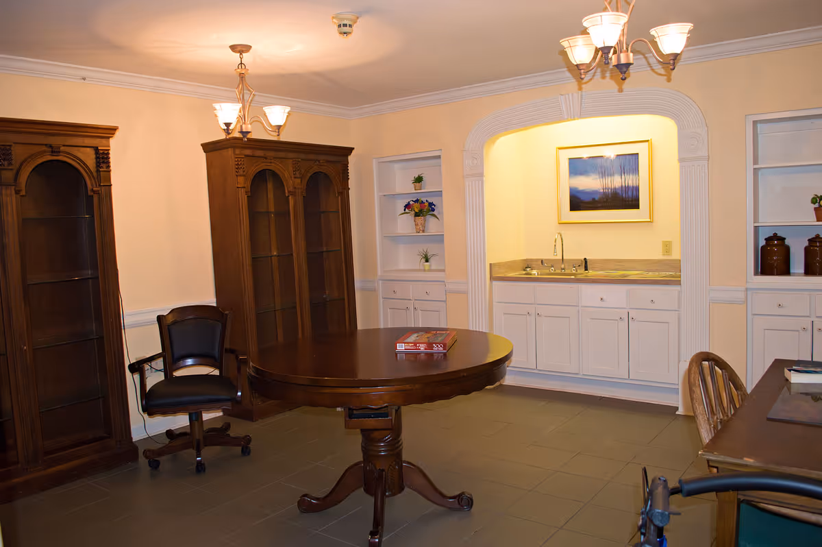 A furnished common room with a round wooden table, chairs, tall display cabinets and a built-in sink alcove.