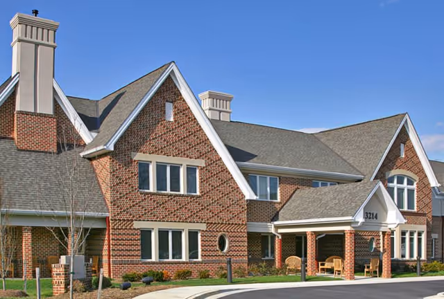 Brick two-story senior living building with pitched roofs, an entrance canopy, and chairs by the driveway.