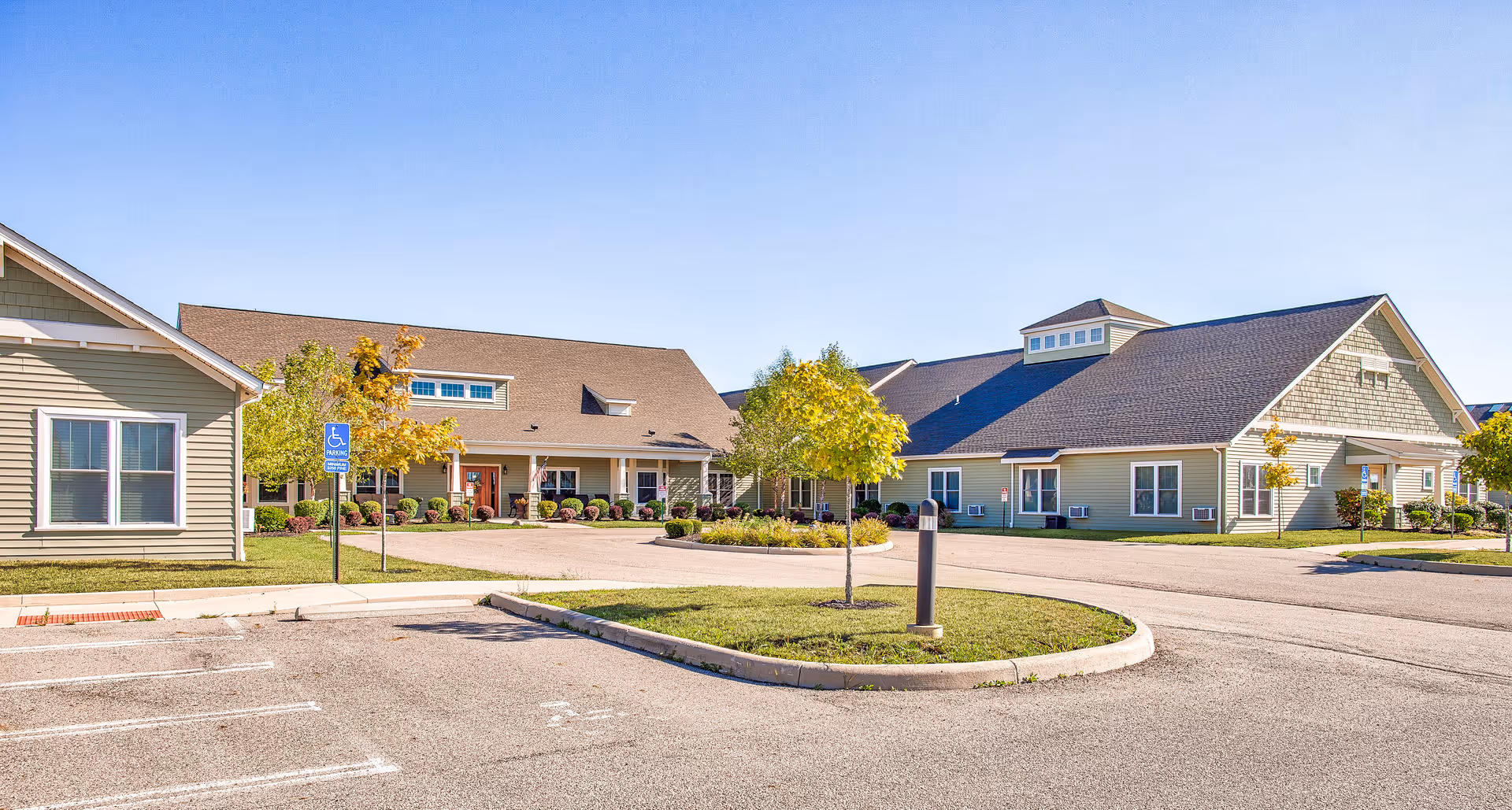 Exterior view of a senior living facility named Landings of Sidney, showing a large building with beige siding, multiple windows, and a sloped roof under a clear blue sky. There is a parking lot with a handicapped parking sign and some small trees and shrubs around the building.