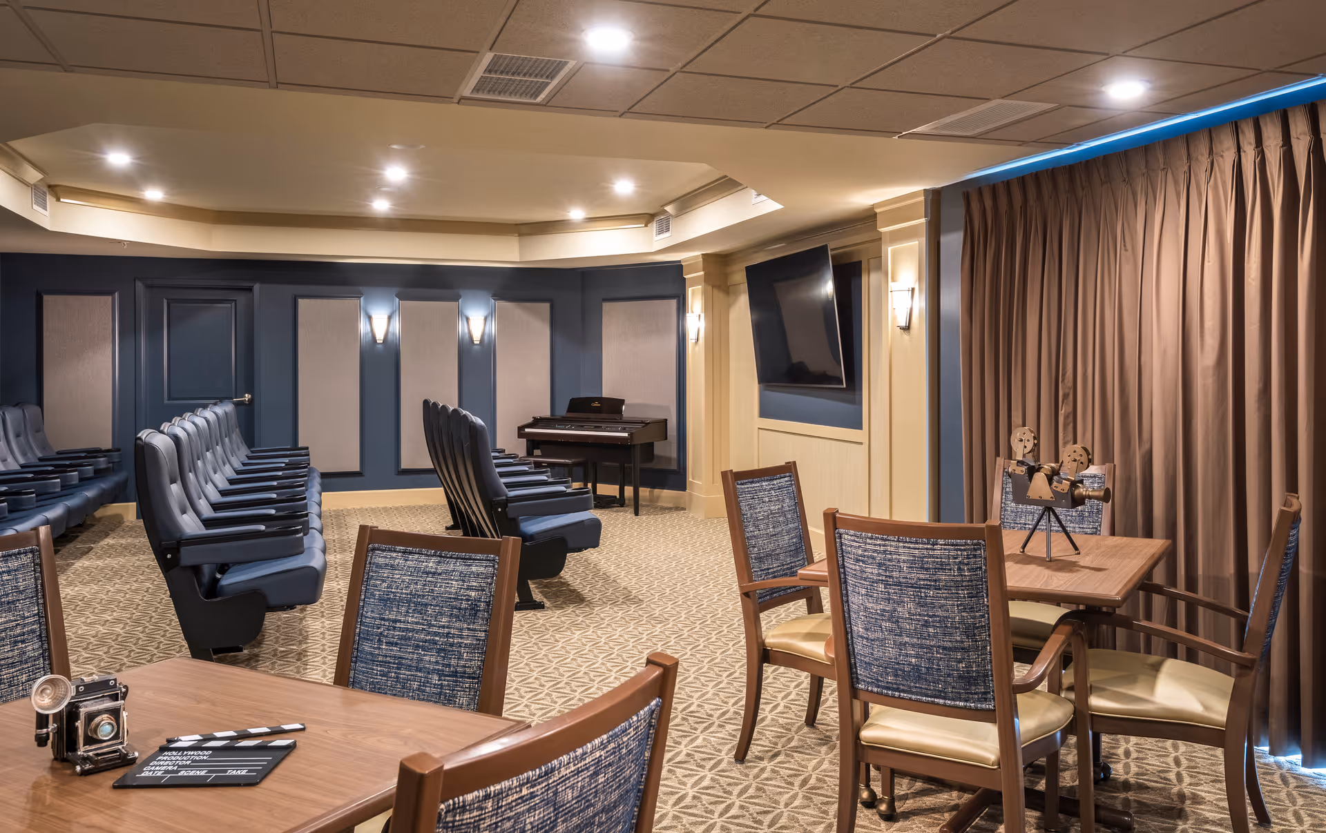 A cozy media room with rows of blue cushioned theater seats facing a wall-mounted flat screen TV. The room has beige patterned carpet, blue and beige walls with wall sconces, and a piano in the corner. In the foreground, there are wooden tables with blue upholstered chairs and decorative items on the tables. Brown curtains cover the windows on the right side.