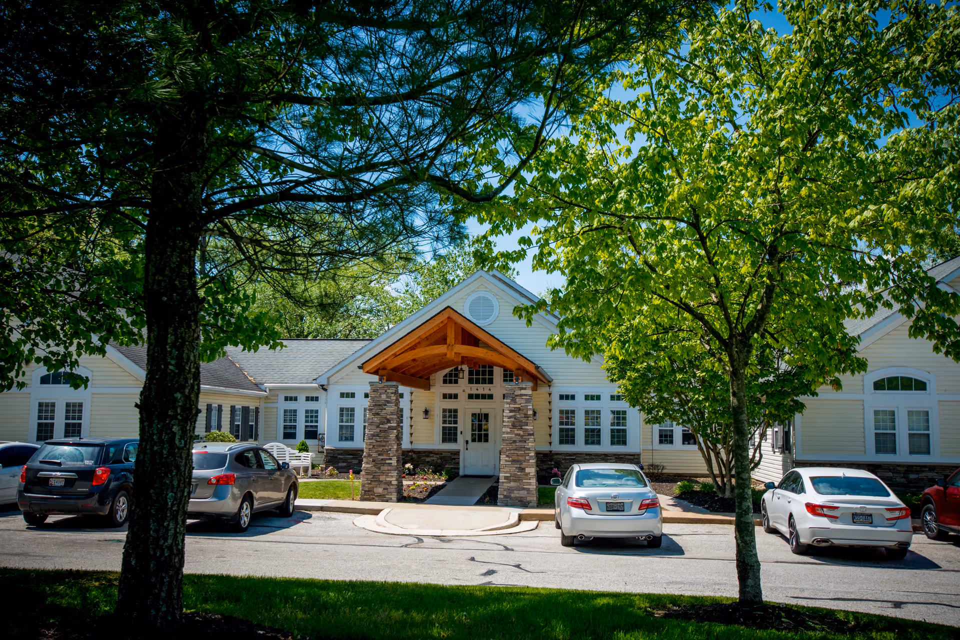 Front exterior view of a single-story building with a peaked roof and stone pillars supporting a wooden awning over the entrance. Several cars are parked in front of the building, and green trees with full foliage frame the scene under a clear blue sky.